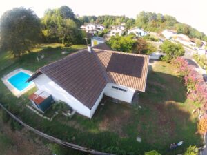 Maison avec panneaux solaires sur le toit, piscine bleue à gauche, jardin et arbres environnants, vue aérienne.